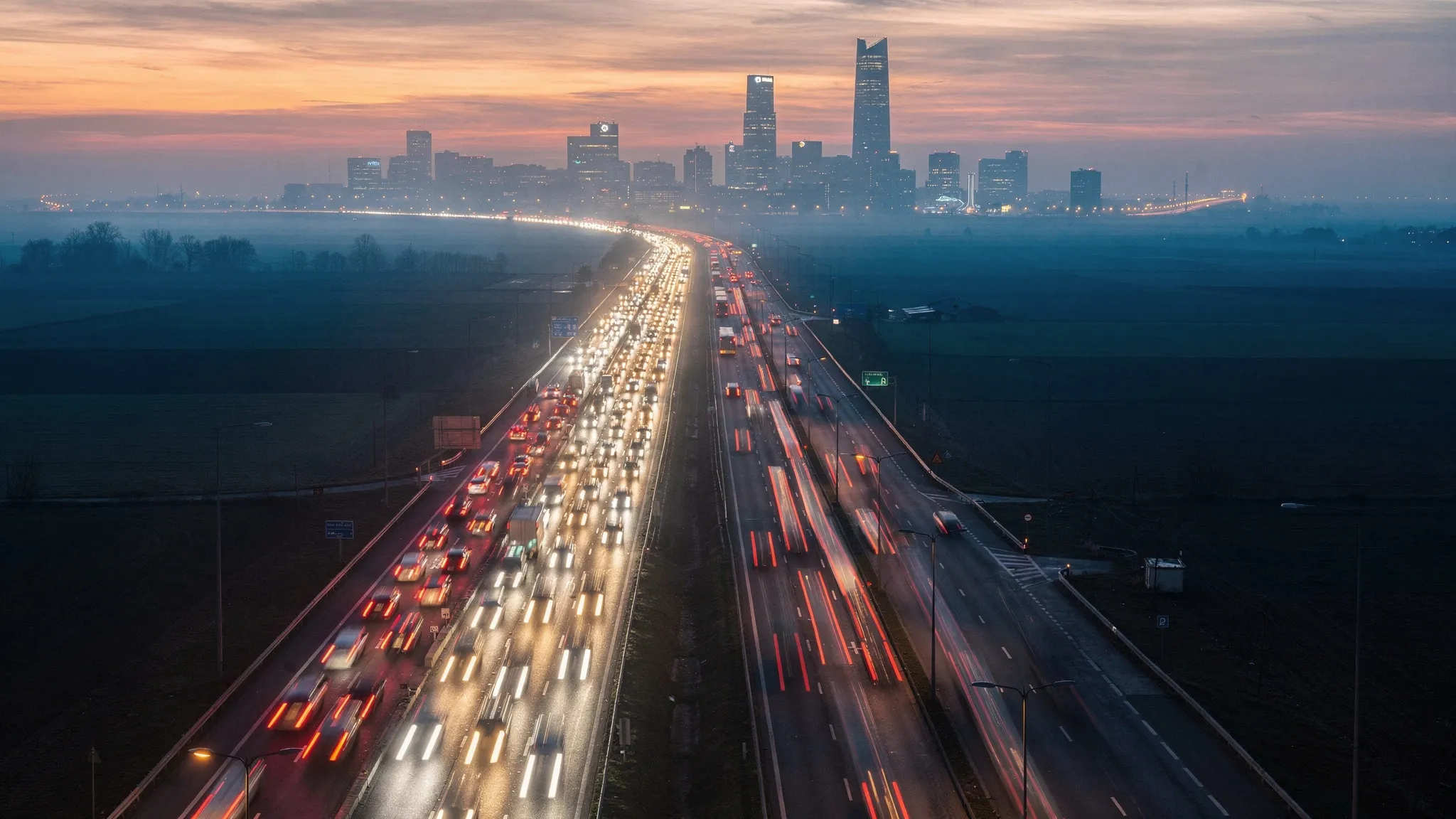 Aerial view at dusk of a highway splitting into two roads toward a distant city, one jammed with cars and headlights, the other carrying fewer, faster-moving cars, in warm amber light.