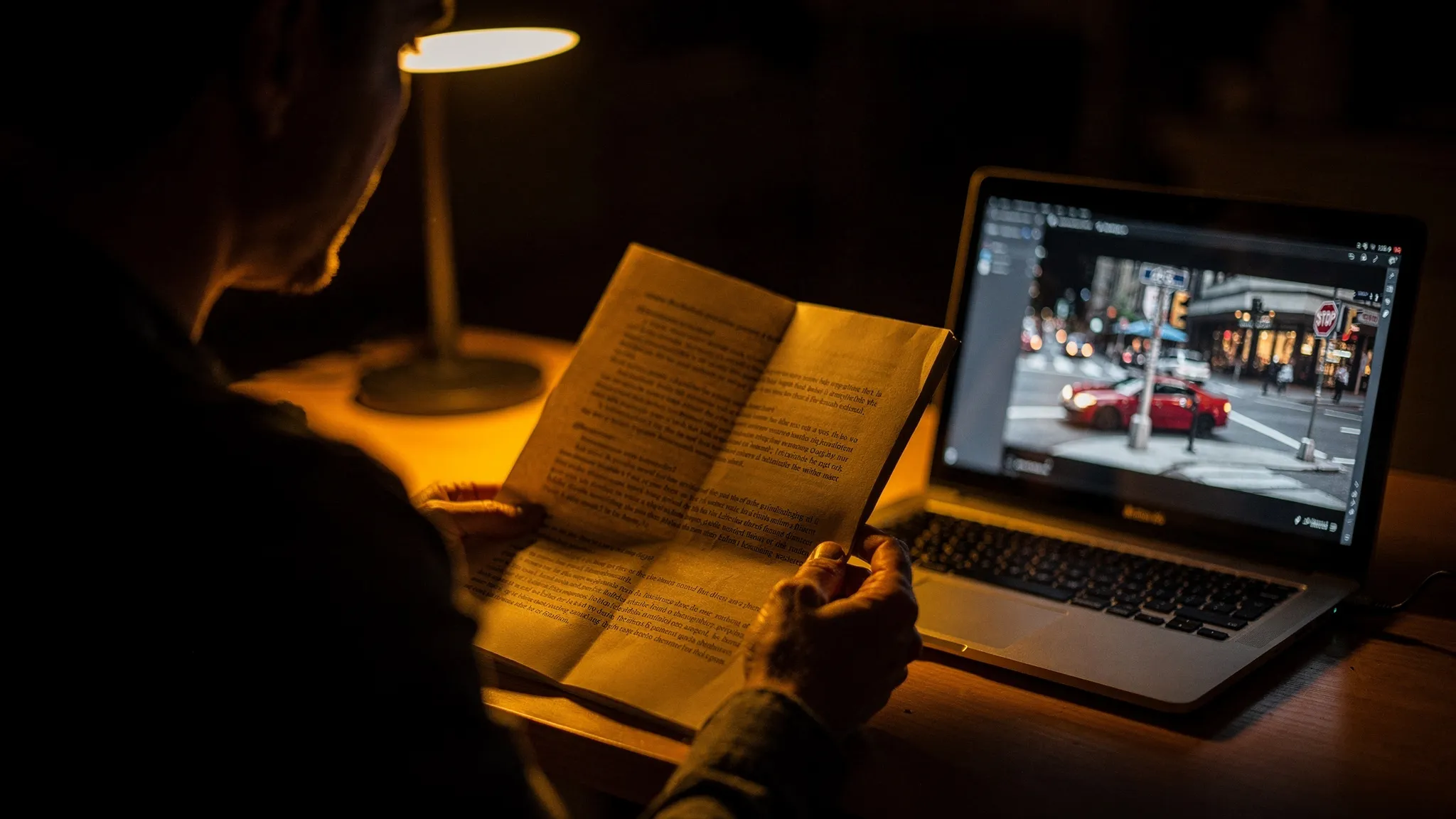 A person reads a printed page under a desk lamp at night, beside a laptop showing a paused street scene with a red car.