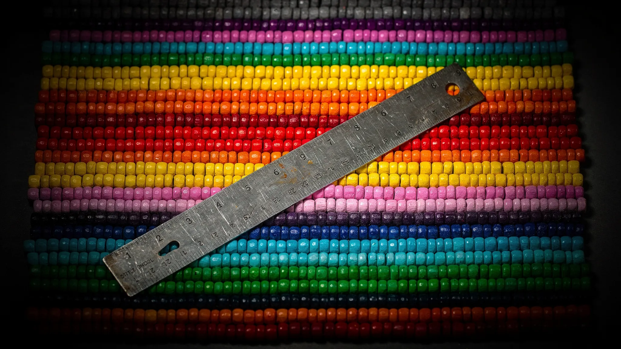 Coloured beads laid out in parallel stripes of different colours on a dark table, a steel ruler across them, cyan rim light.
