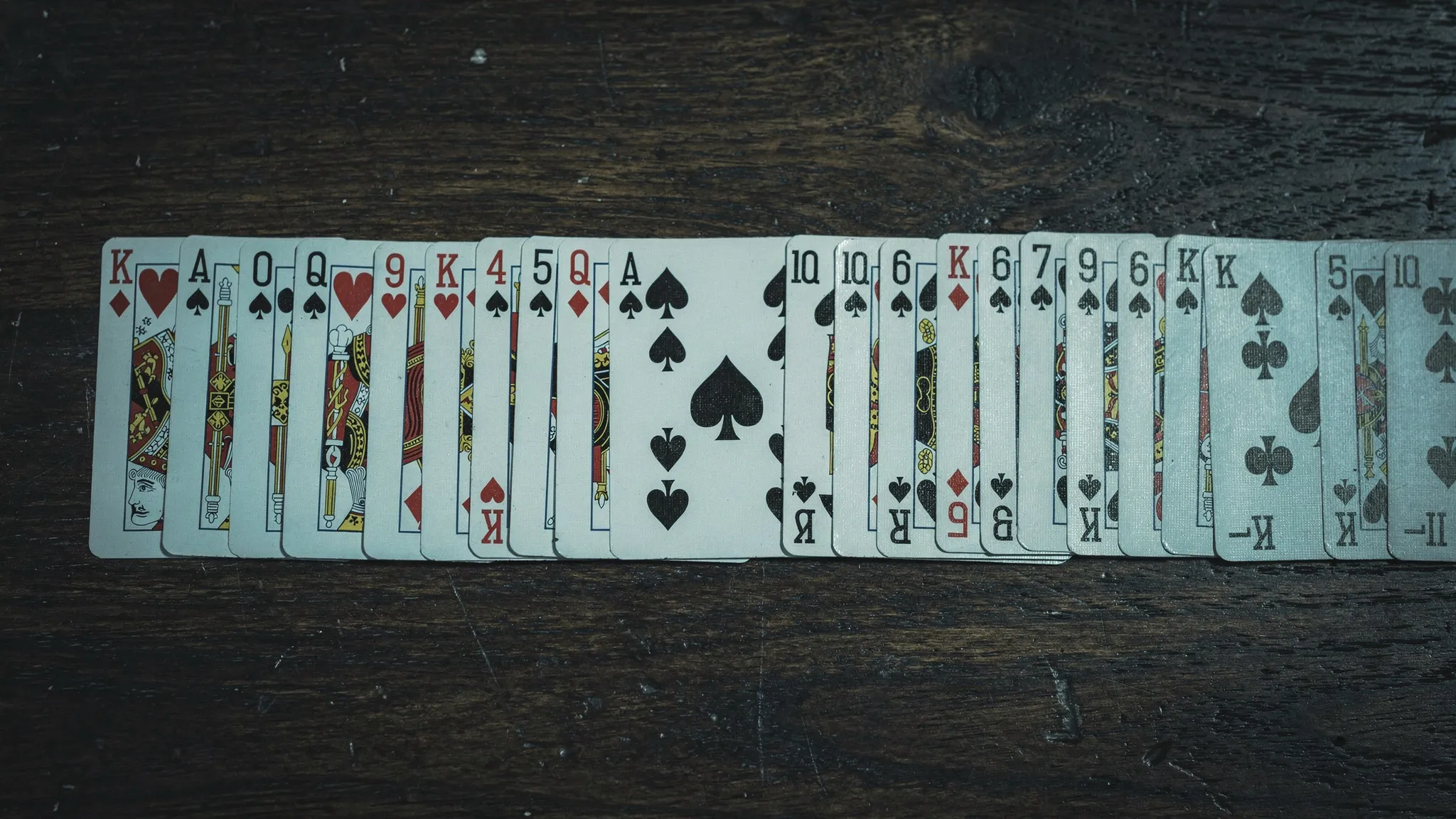 Playing cards dealt face up in a long row on a dark wooden table, an ace of spades appearing among the first cards.