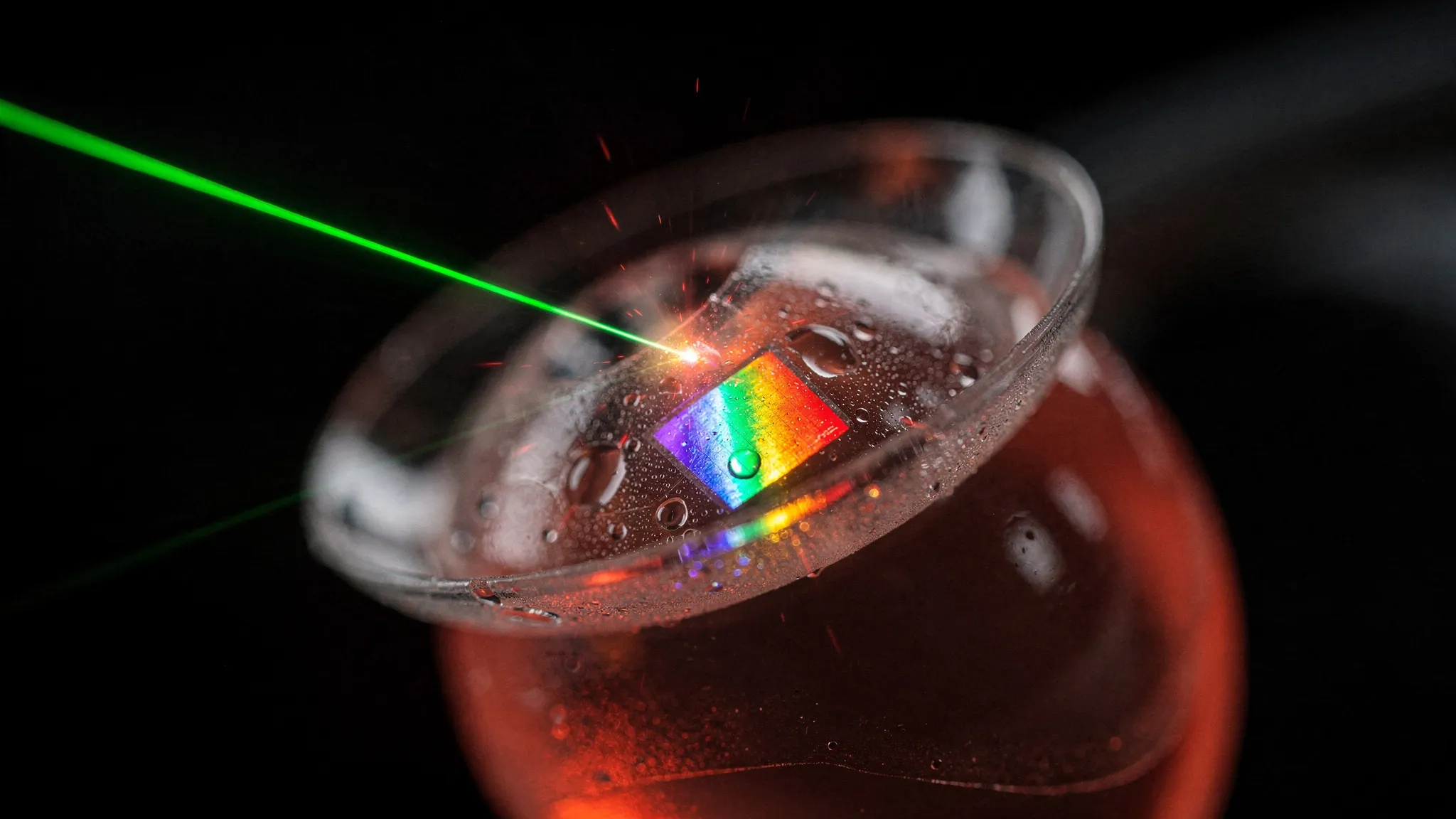 A clear soft contact lens resting on a red-lit artificial eye, a green laser beam hitting a small rainbow-sheened patch near its centre.