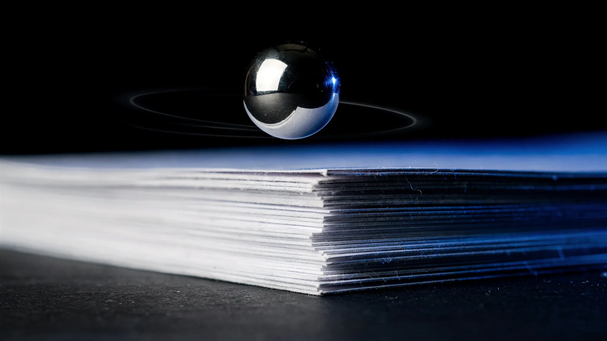 A small steel ball bouncing above a neat stack of white printer paper on a dark table, lit in cobalt blue, with faint circular motion trails around it.