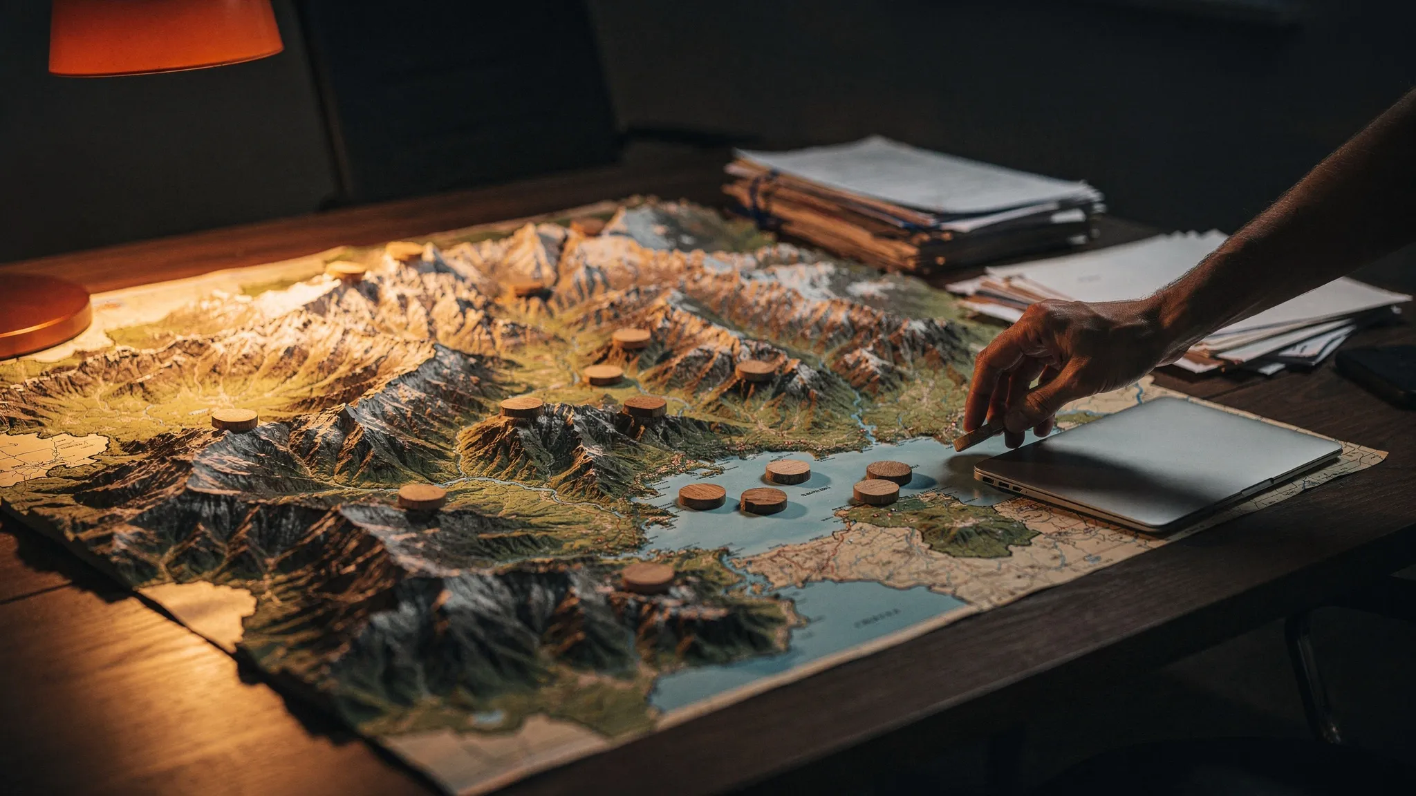 A relief map of Switzerland on a desk, small markers placed on several cantons, a placement officer's hand beside a laptop, in amber light.