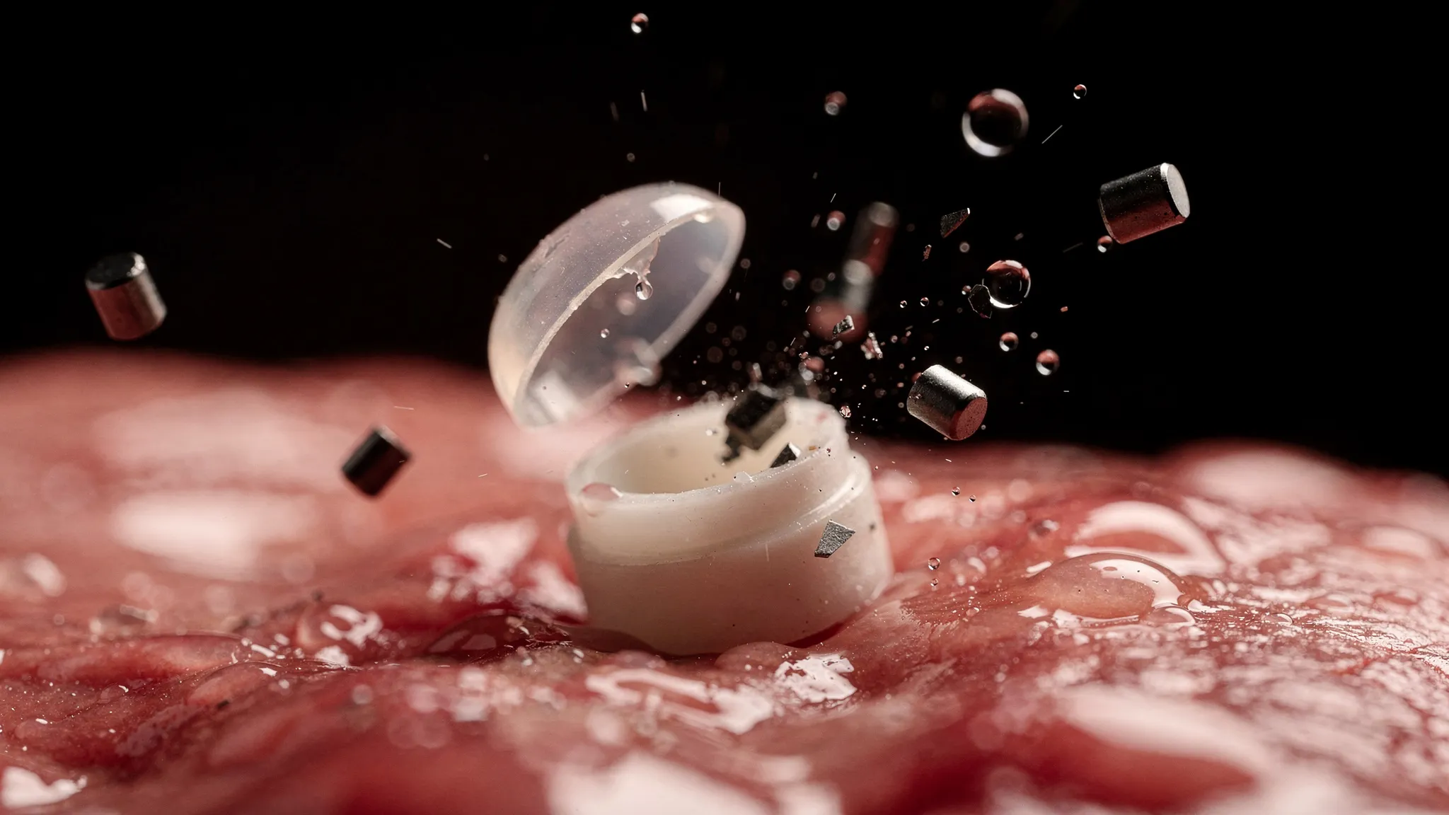 A tiny capsule bursting open on stomach tissue, its soft transparent dome flipping off and small dark magnetic pieces flying out, red light.